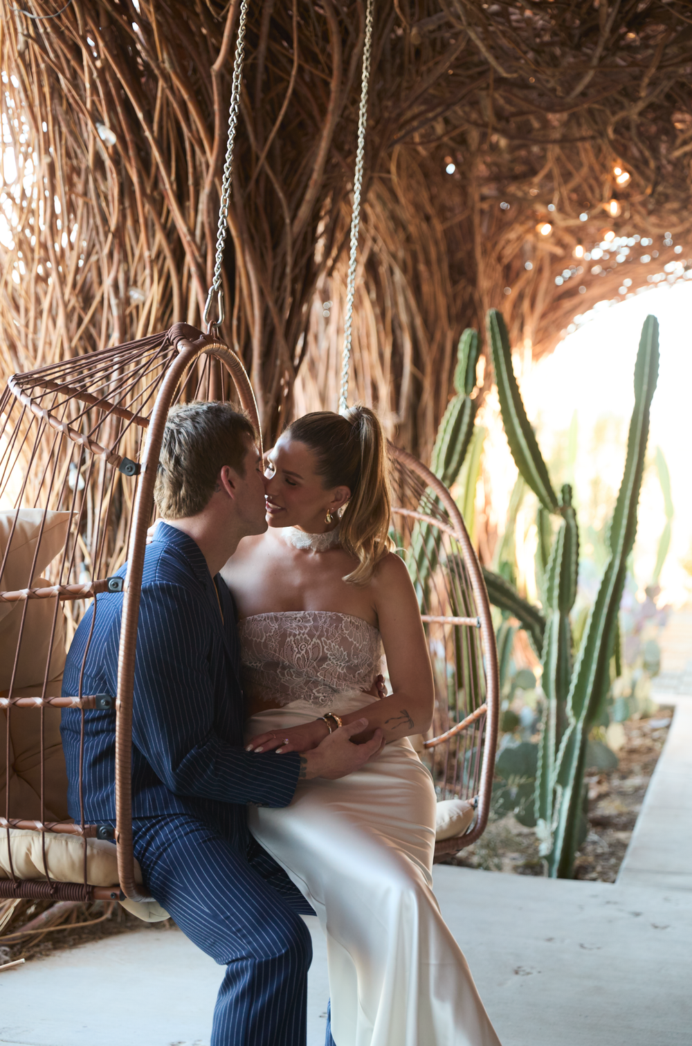 Bride and groom share a romantic moment on a rattan swing chair surrounded by cacti and woven decor.