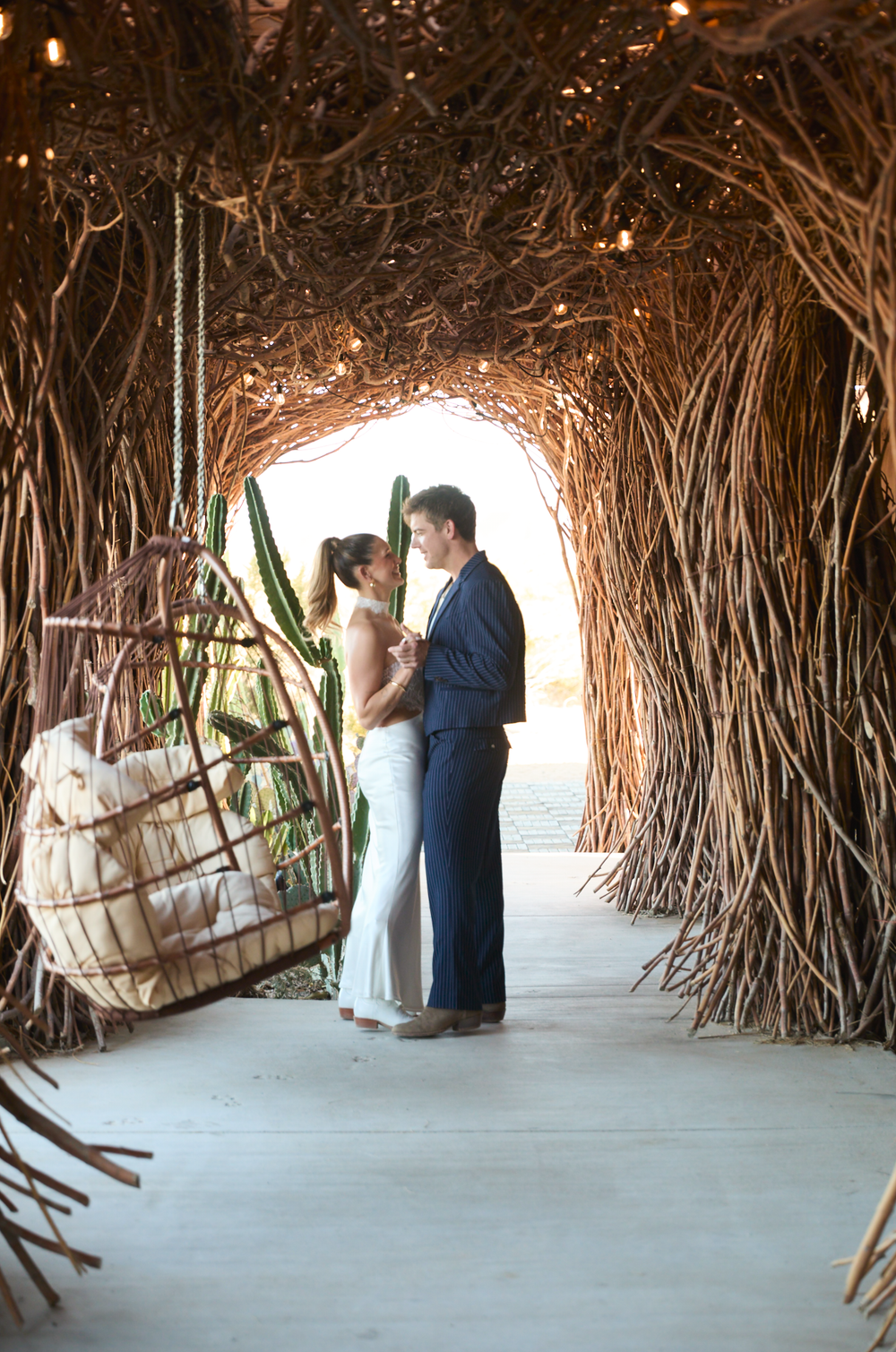 Couple shares romantic moment in rustic twig tunnel with hanging wicker chair and cacti at bohemian wedding venue.