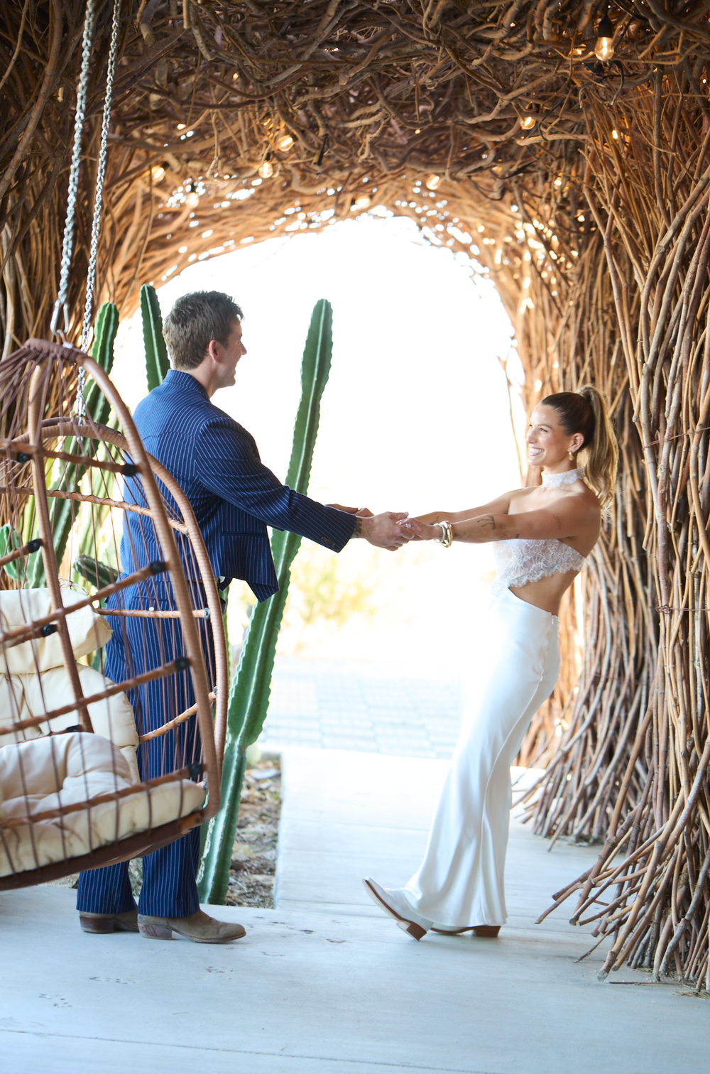Couple holding hands in a rustic woven branch tunnel with cactus, bride in white gown, groom in blue suit.
