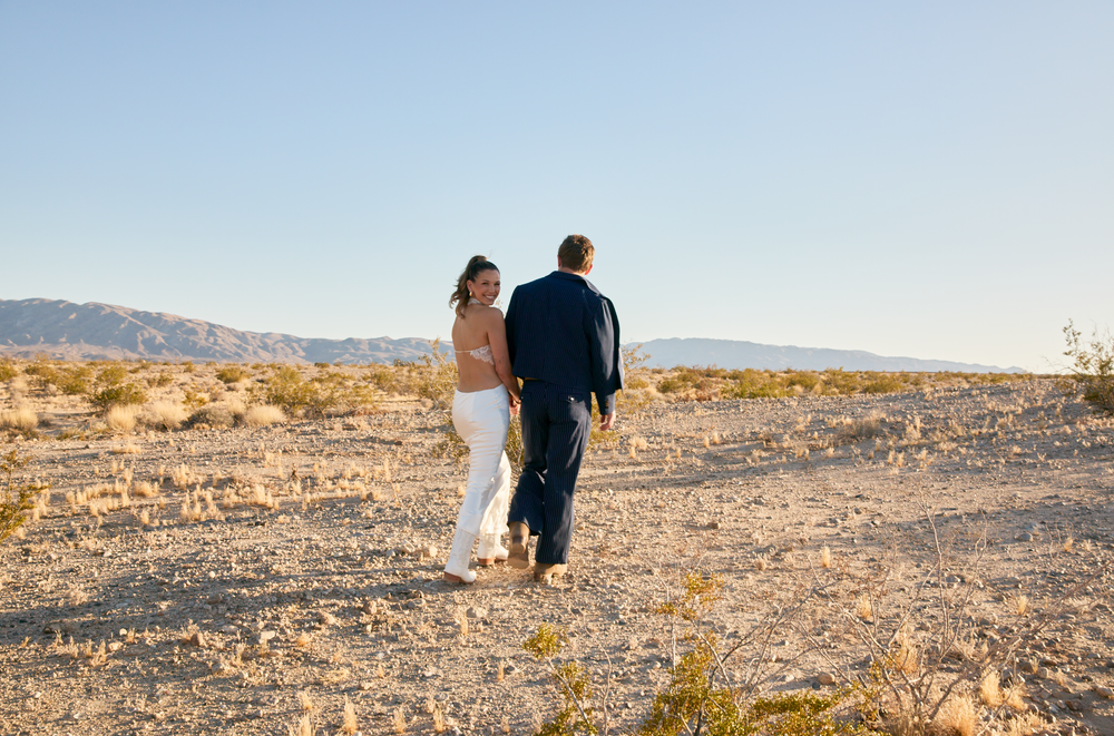 A couple in wedding attire walks hand-in-hand through a desert landscape with mountains in the background.