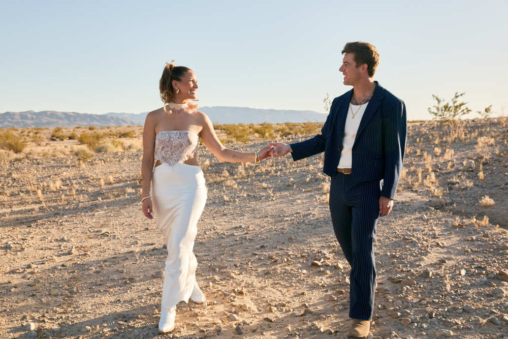 A couple holds hands and dances in a sunny desert landscape, dressed in elegant white and navy blue outfits.