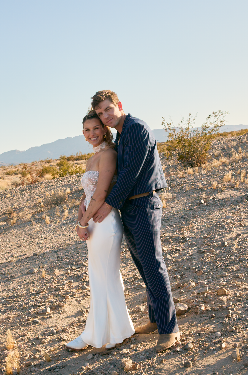 Couple in wedding attire embracing in desert landscape, bride in white gown, groom in navy suit, mountains background.