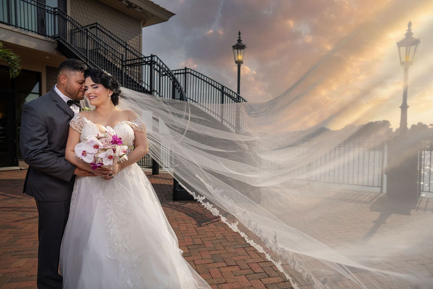 Bride and groom embrace at sunset, her long veil flowing dramatically in the wind at a waterfront wedding venue.