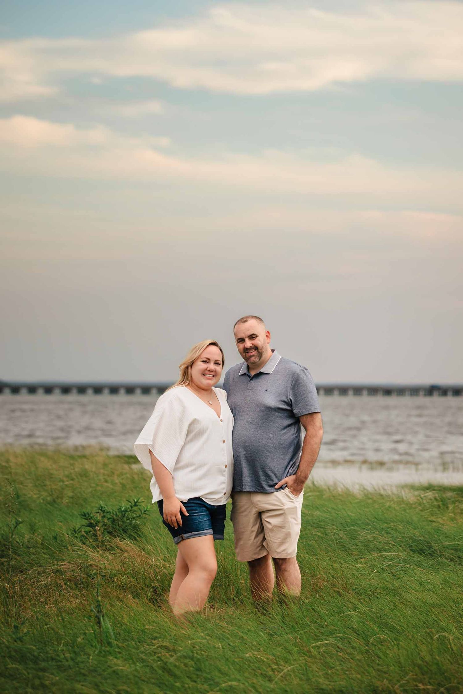 A smiling couple stands together on a grassy waterfront at sunset, woman in white shirt and denim shorts, man in gray polo.