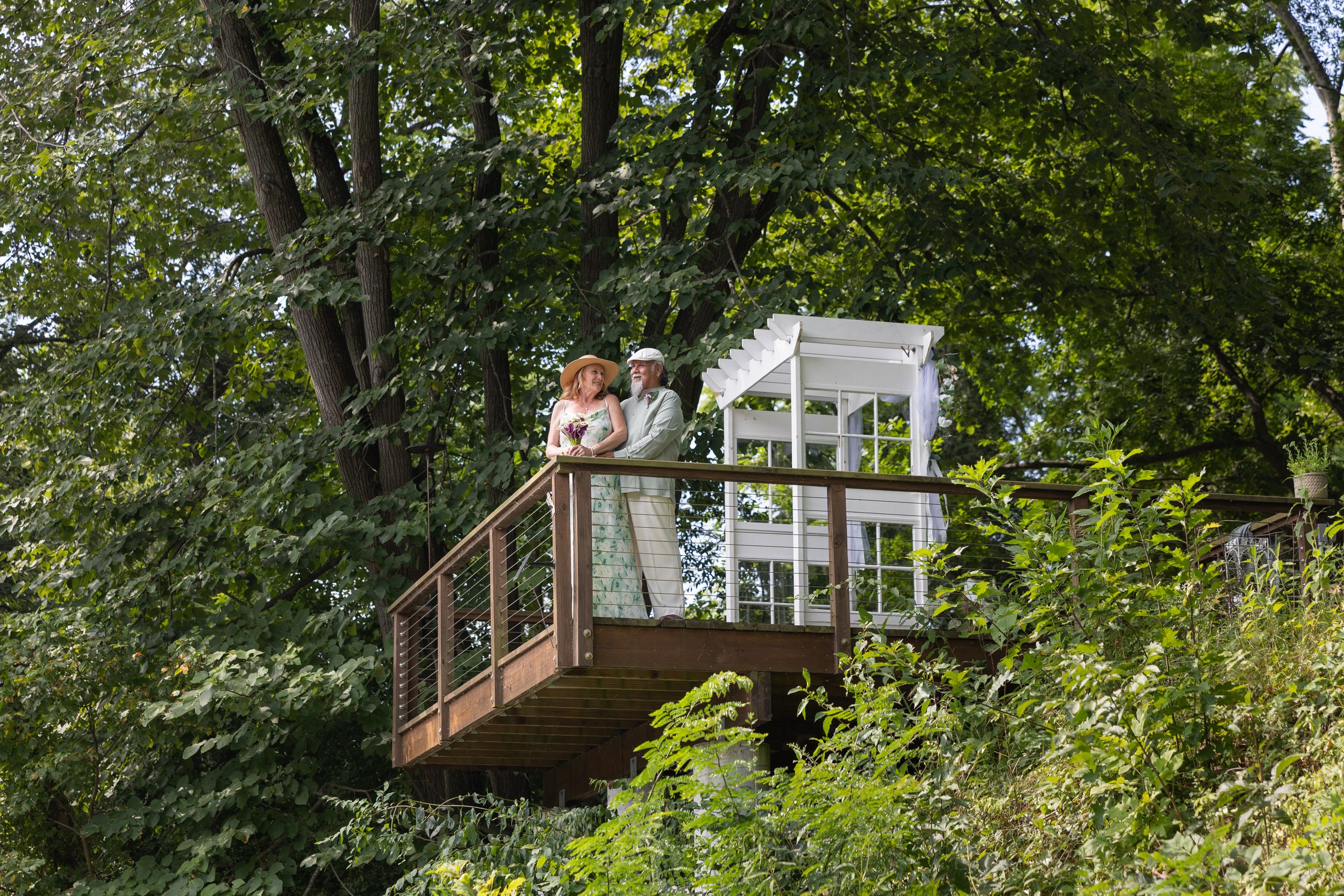 Two people in vintage attire stand on a wooden deck surrounded by lush green trees near a white gazebo.