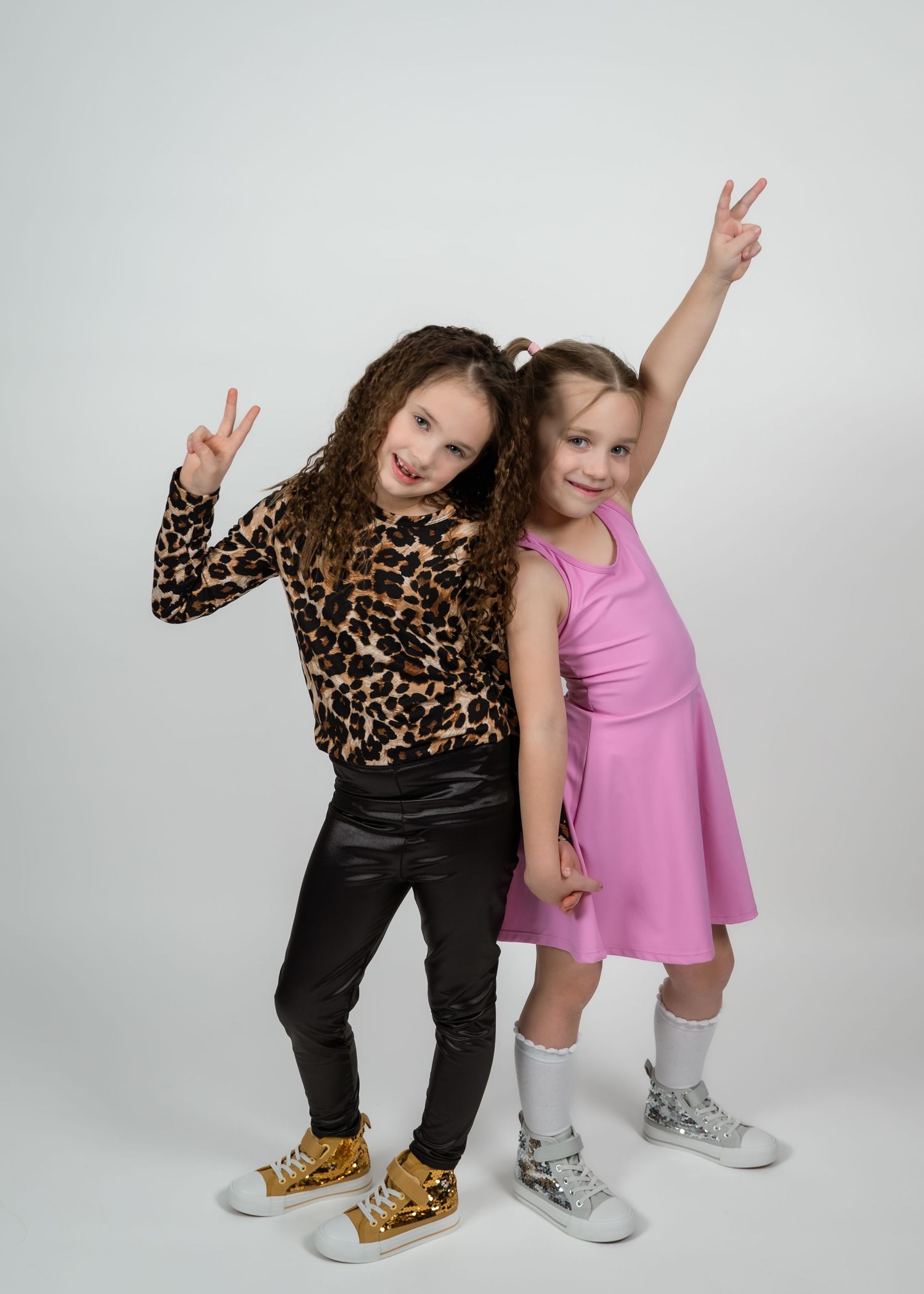 Two young girls posing playfully, one in leopard print top with black pants, other in pink dress, both making peace signs.