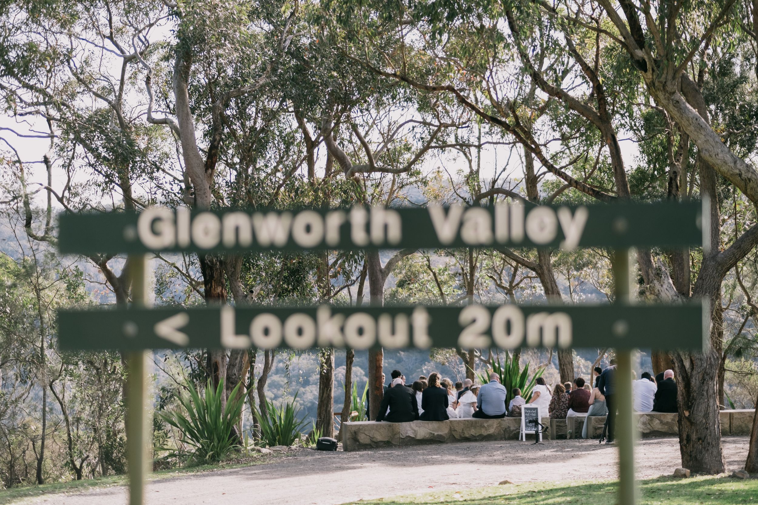 Glenworth Valley Lookout sign with wedding guests gathered at a scenic bush lookout 20 metres away.