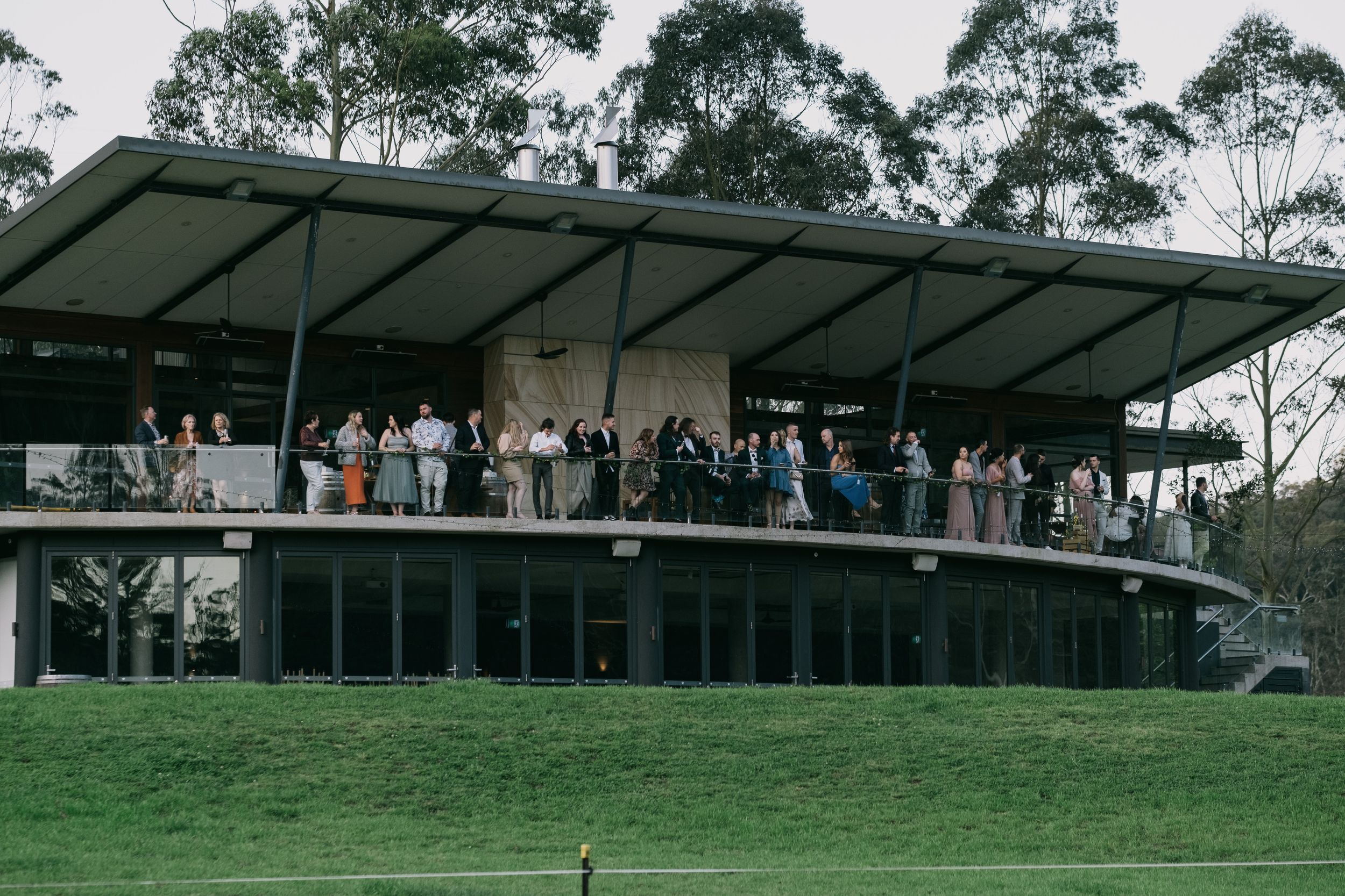 Wedding guests gather on a modern curved pavilion overlooking a lush green lawn in bushland.