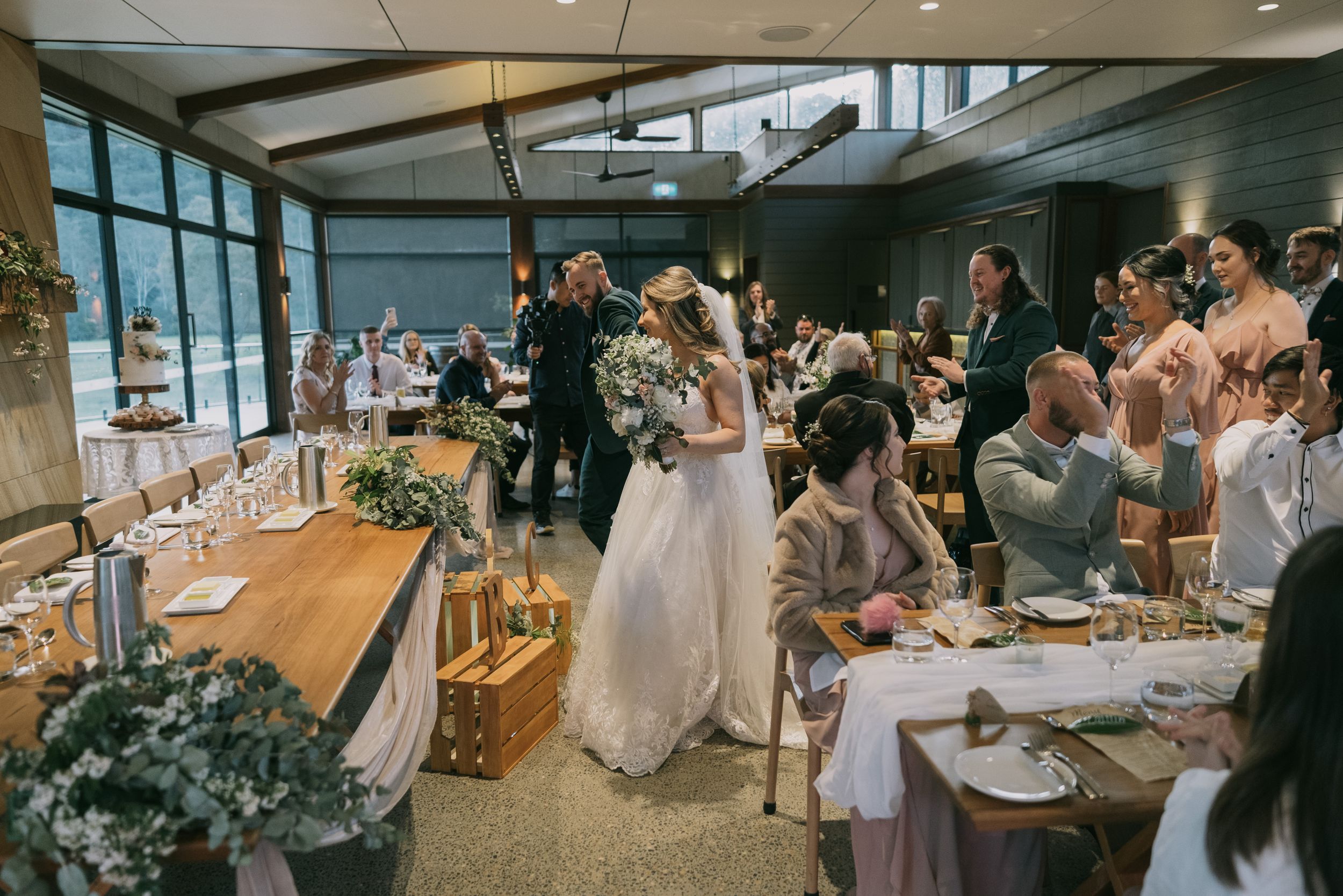 Bride enters wedding reception hall in a flowing white gown as guests applaud and celebrate.