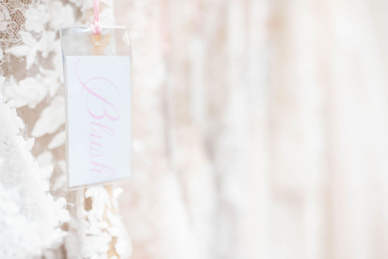 A delicate white bridal tag hangs among soft ivory wedding dresses in a bridal boutique.