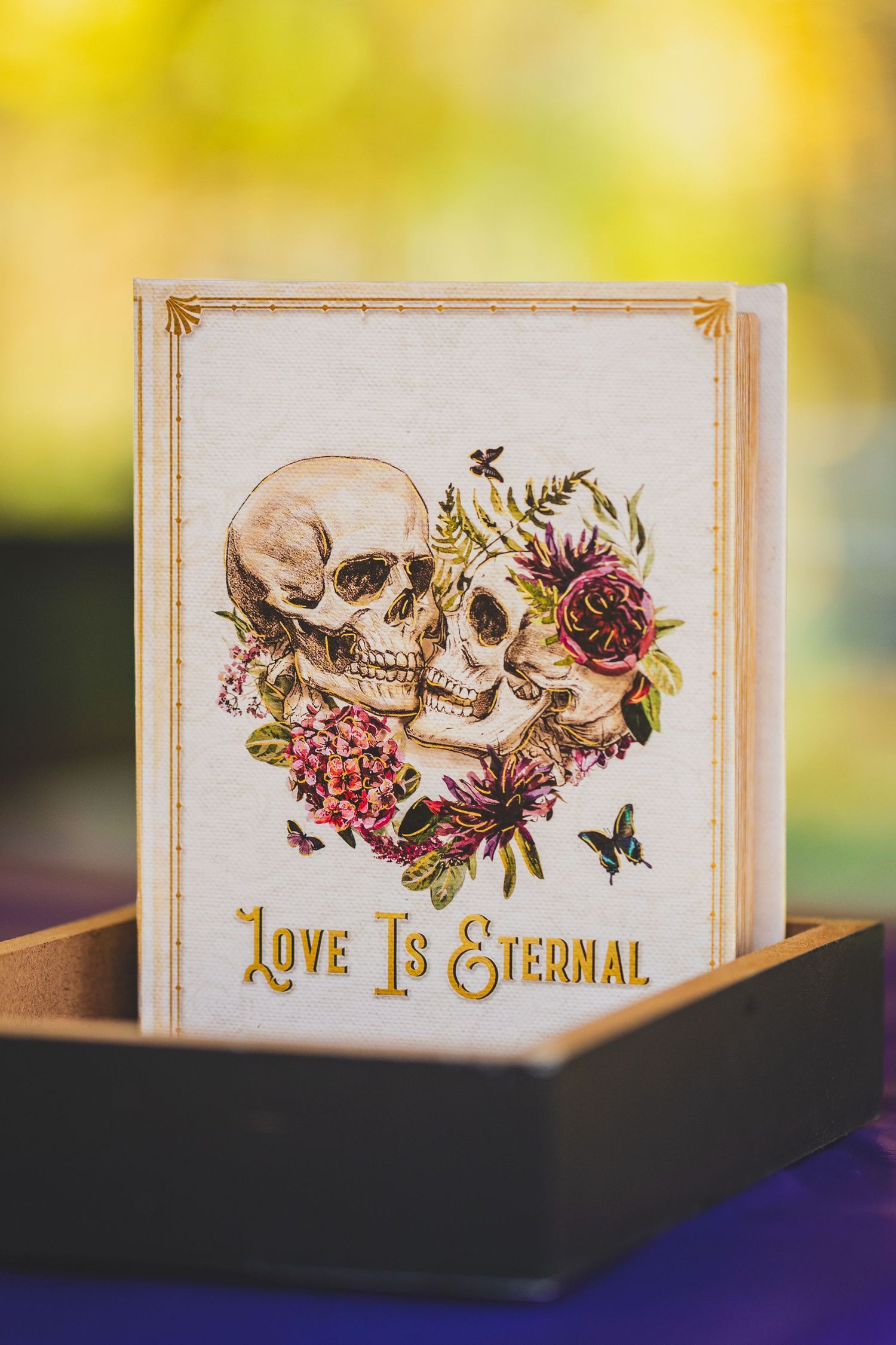 Gothic wedding guest book with two skulls and flowers, reading Love Is Eternal, displayed in a wooden box.