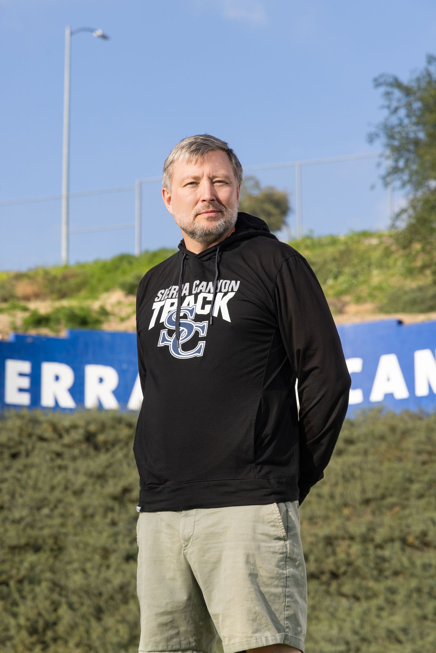 Man in black Sierra Canyon Track hoodie and khaki shorts stands outdoors near blue banners and green shrubs.