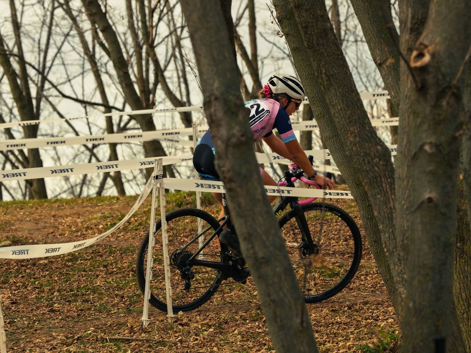A cyclist in a pink and blue kit races a cyclocross bike with pink bar tape past bare trees and branded course tape at a Wisconsin race