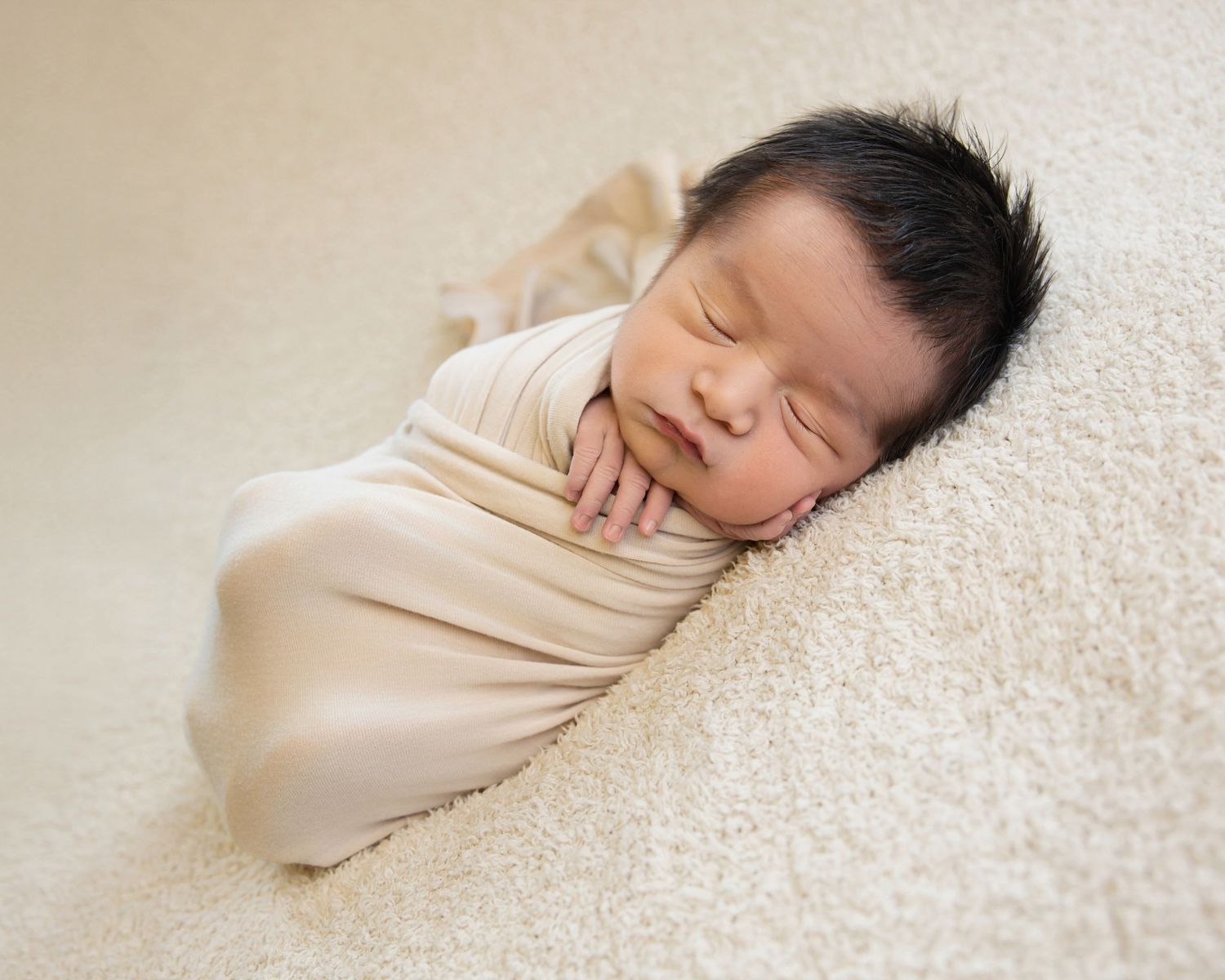 Sleeping newborn with dark hair wrapped in cream fabric and resting on a soft neutral blanket.