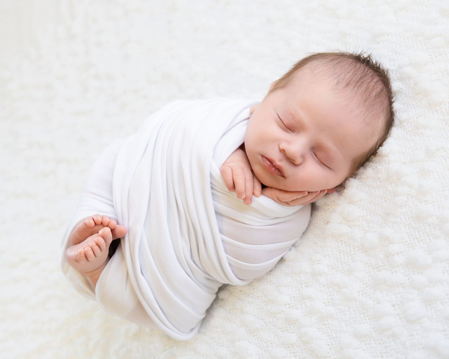 Peaceful newborn baby swaddled in white wrap sleeping on soft fluffy white blanket.