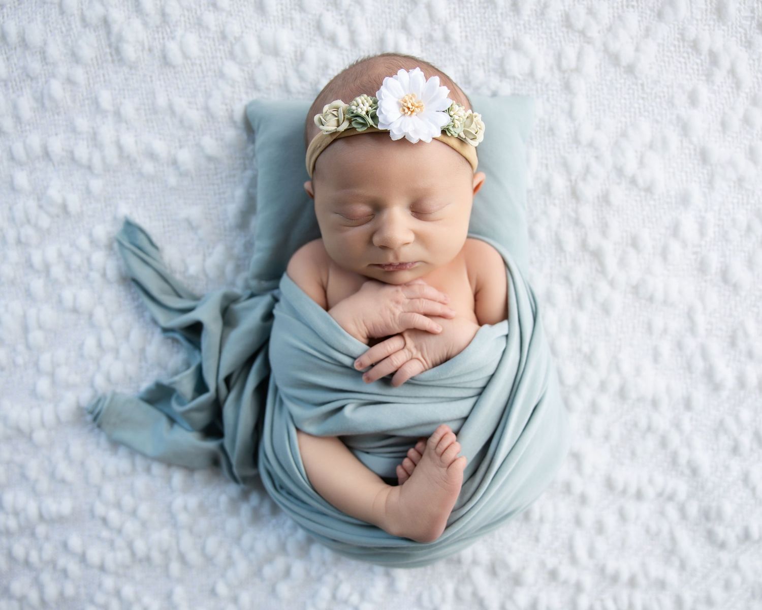 Newborn baby girl sleeping peacefully wrapped in a soft teal swaddle with a delicate daisy headband on white background.