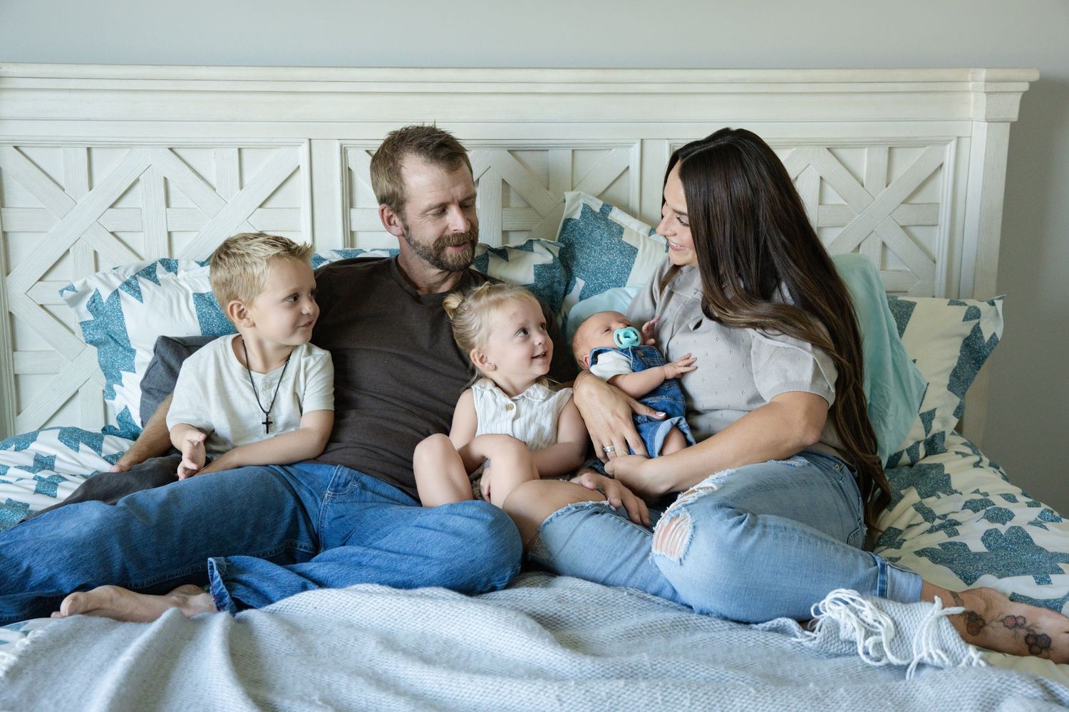Happy family with two toddlers and a newborn baby relaxing together on a bed with blue patterned bedding.
