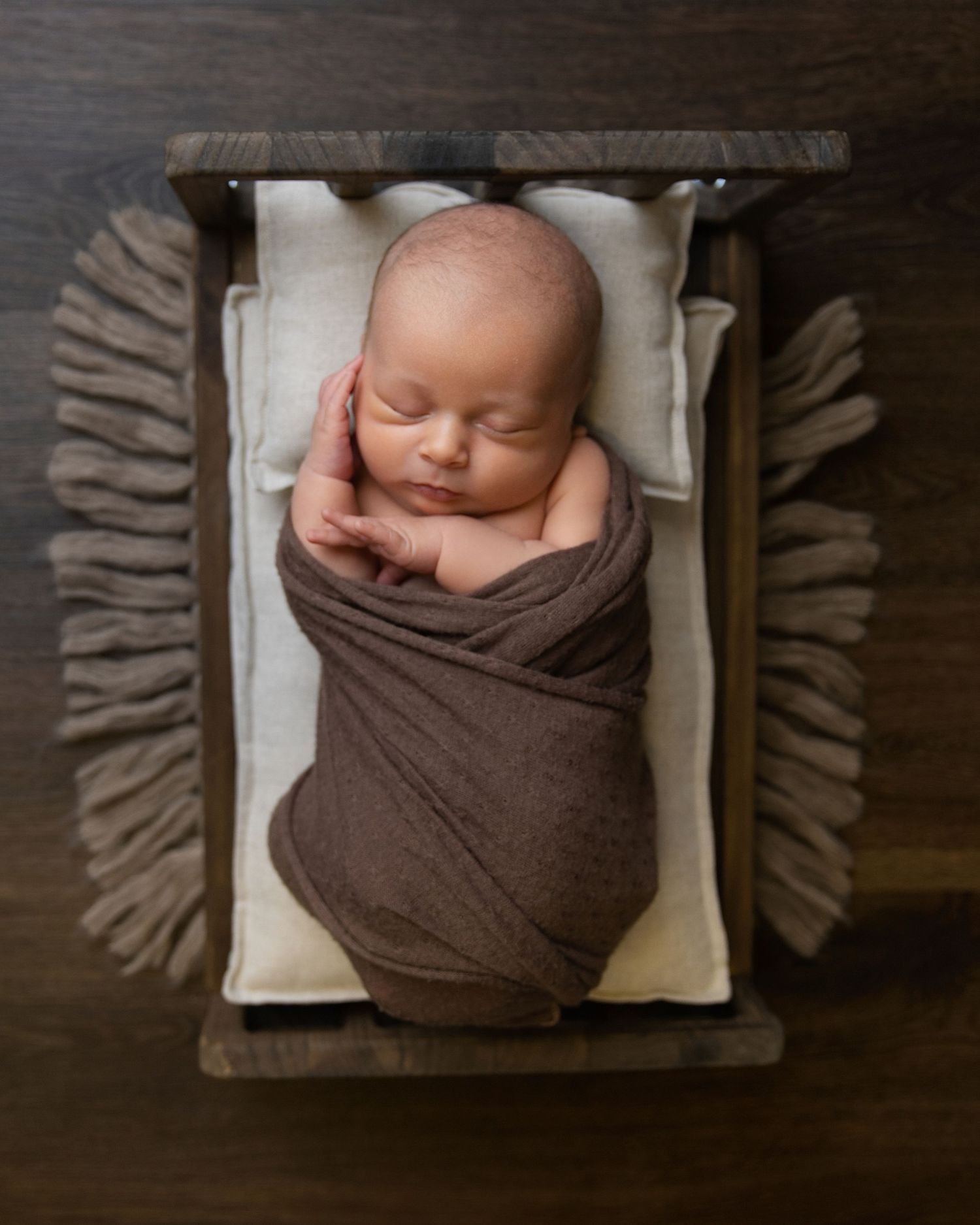 Newborn baby swaddled in brown wrap sleeping peacefully in a rustic wooden crib with cream lining.