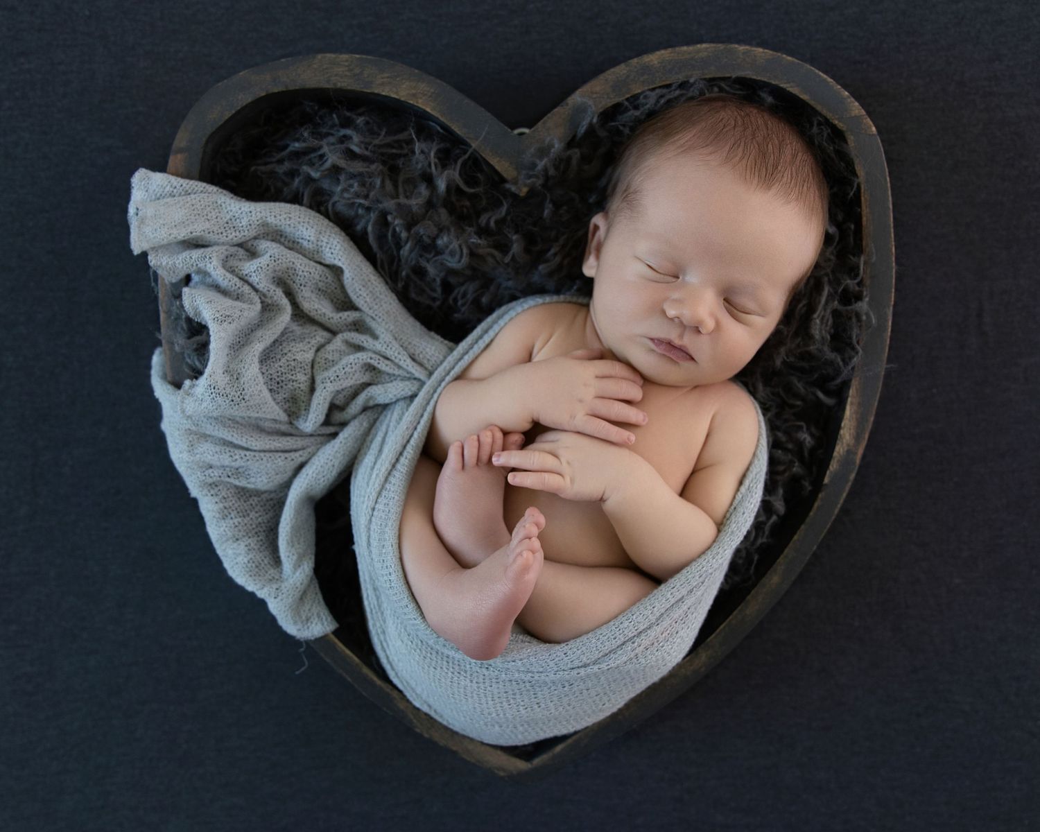 Sleeping newborn baby wrapped in gray cloth inside a wooden heart-shaped bowl on dark background.
