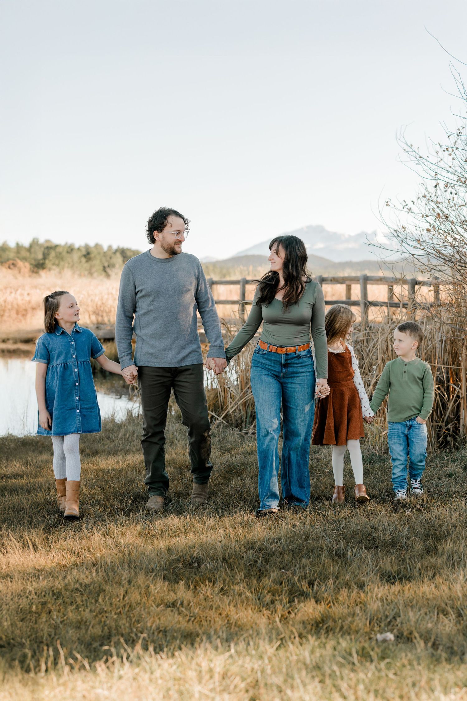Family of four holding hands in a field near a lake with mountains in the background on a clear day.