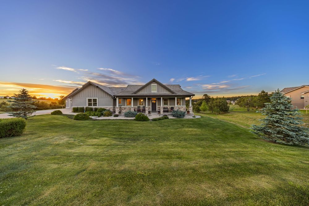 Ranch-style home with covered porch, lush green lawn, mature trees, and stunning sunset sky in a serene rural setting.