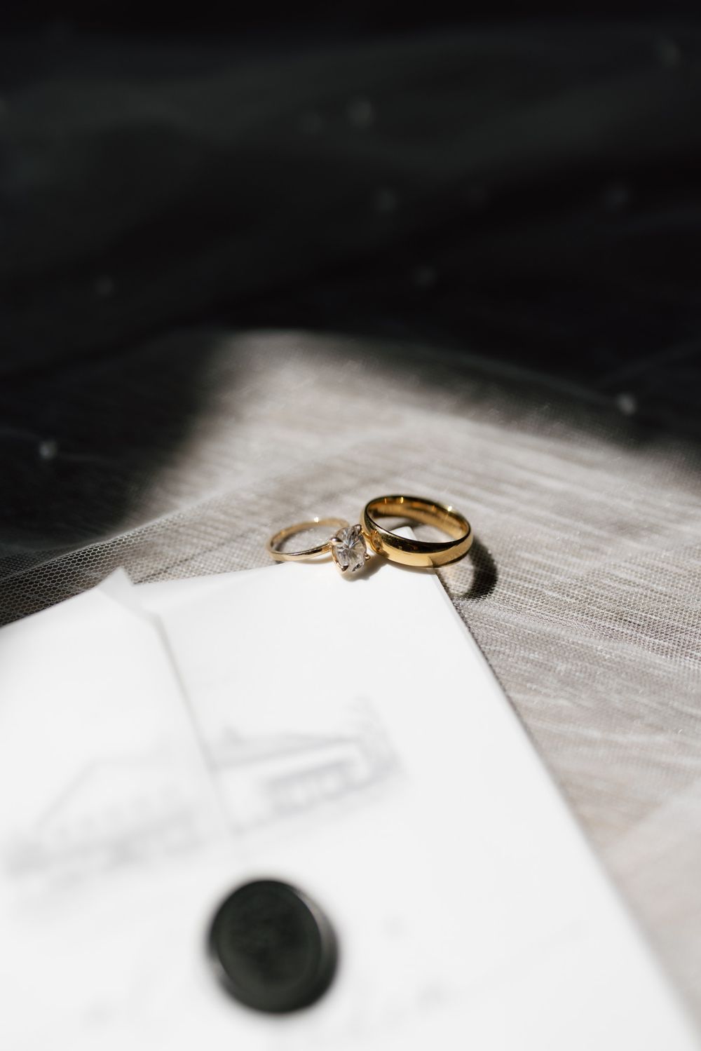 Gold wedding band and diamond engagement ring resting on white envelope with wax seal and sheer veil.
