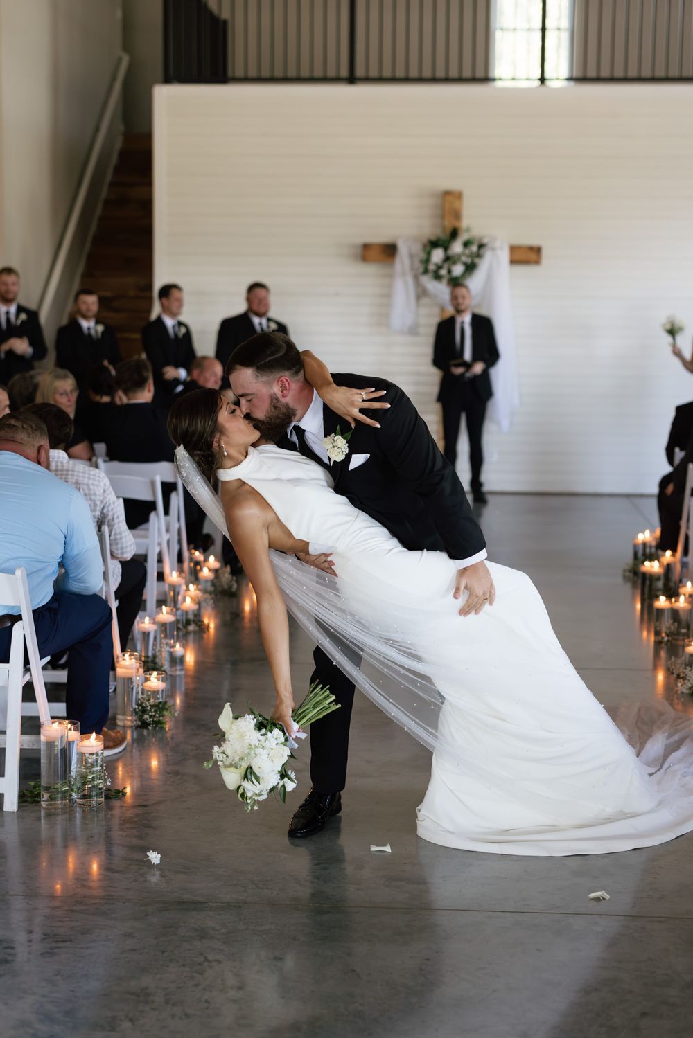 Groom dips bride for a romantic kiss at wedding ceremony aisle lined with candles, guests seated nearby.