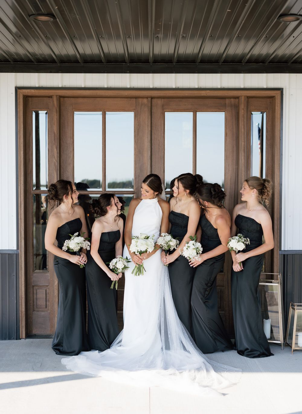 Bride in white gown with bridesmaids in black strapless dresses holding white bouquets outside wooden doors.