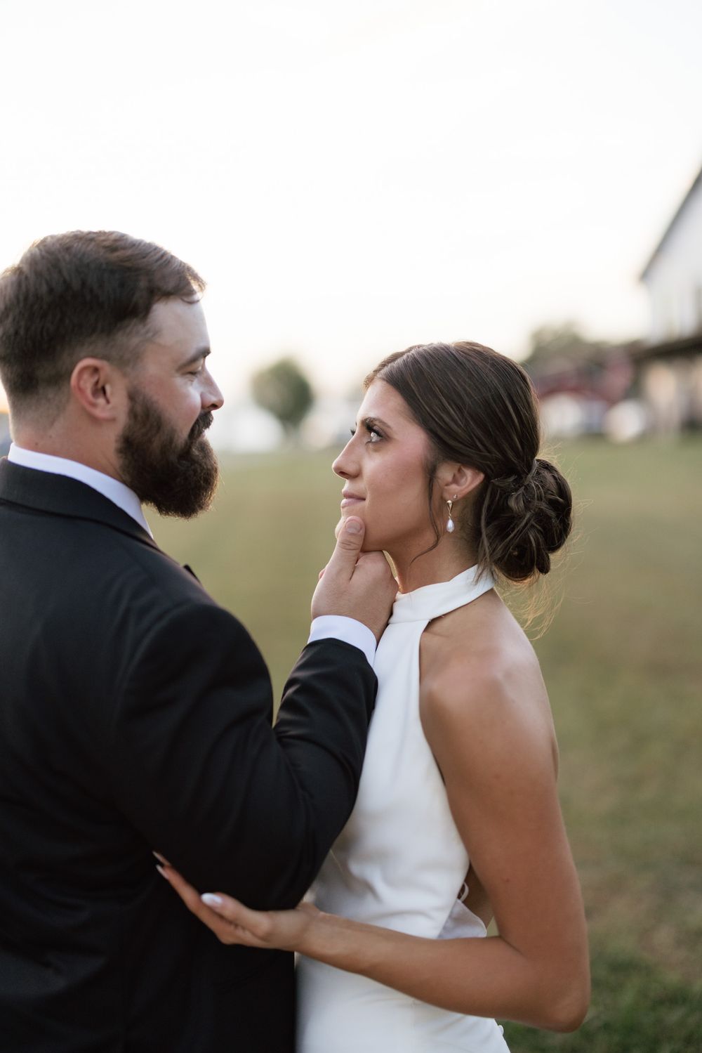 Bearded groom in black suit tenderly holds brides face as she gazes up at him during outdoor wedding portrait.