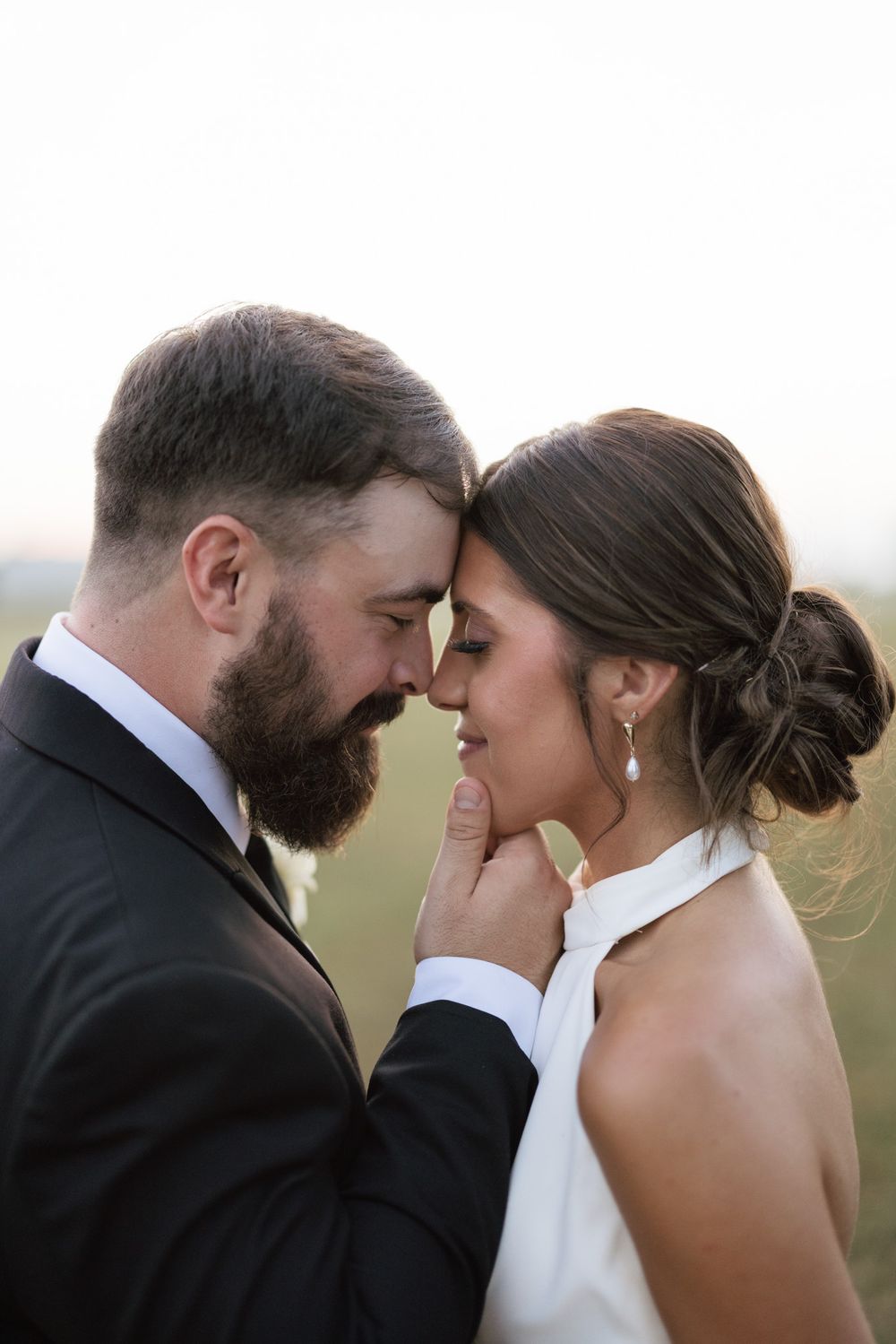 Bearded groom in black suit tenderly holds bride's chin as they lean foreheads together in romantic outdoor wedding portrait.