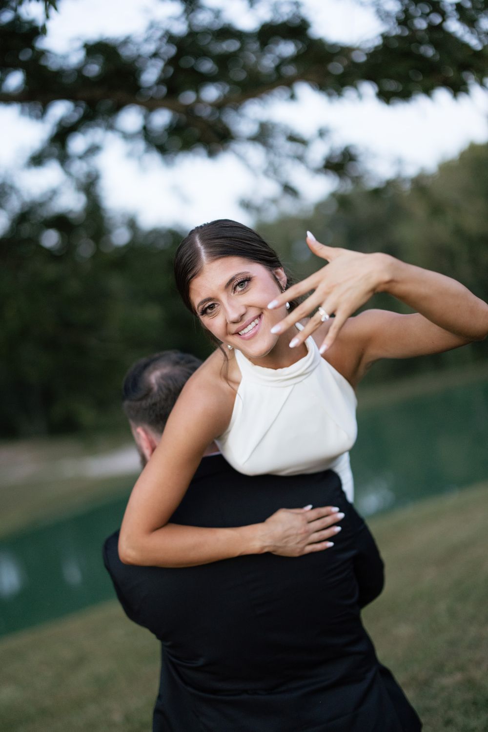 Smiling bride in white halter dress shows off engagement ring while hugging groom outdoors near a pond.