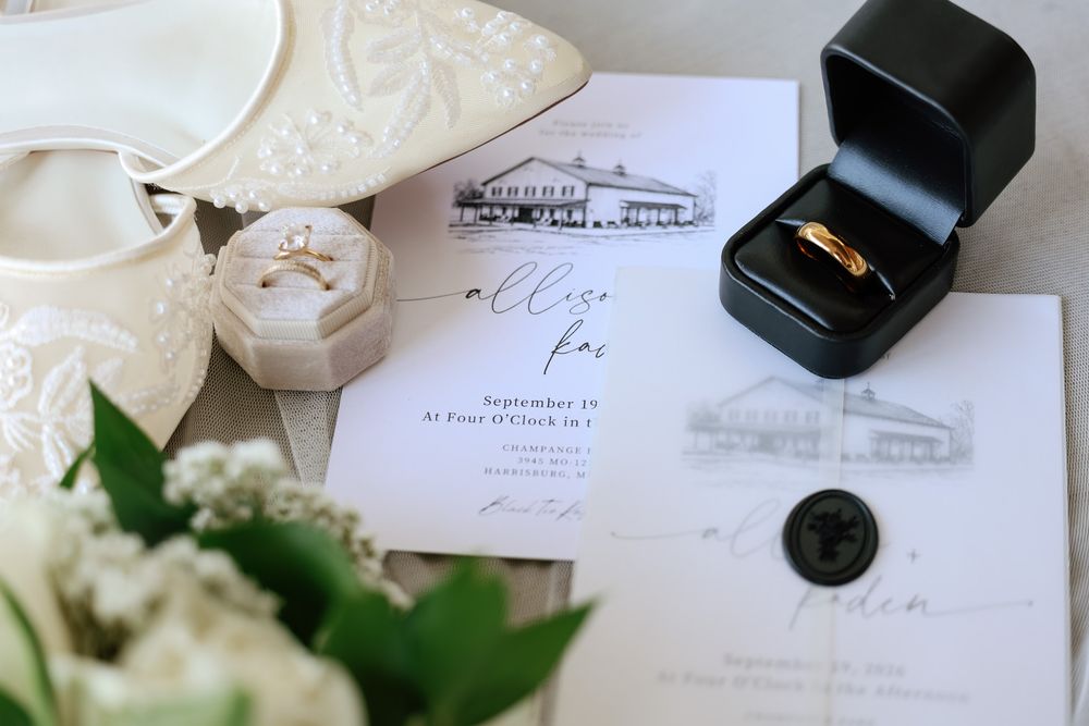 Elegant wedding flat lay featuring gold bands in a black ring box, floral invitations, wax seal, and bridal shoes.