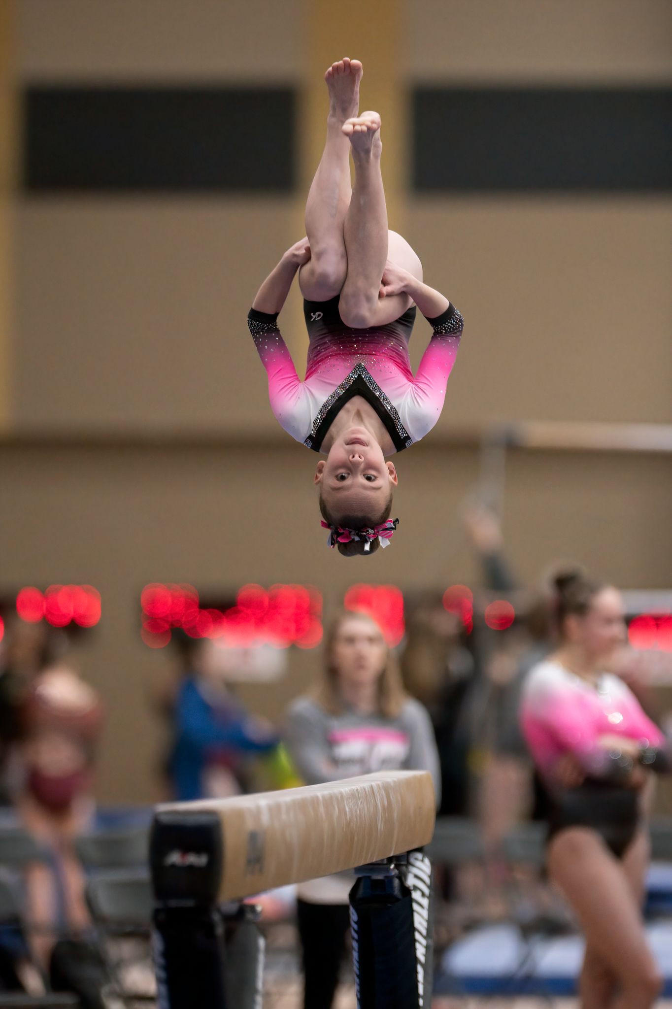 Gymnast in pink leotard performs aerial flip over balance beam during competition.