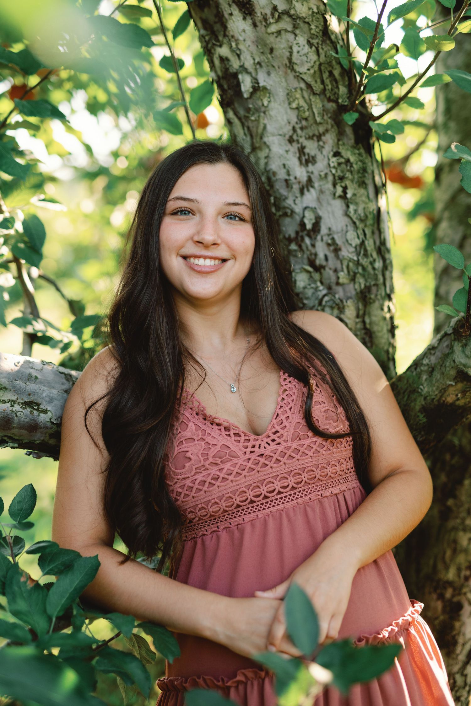 Young woman with long dark hair smiles outdoors, leaning against a tree in a pink dress surrounded by green leaves.