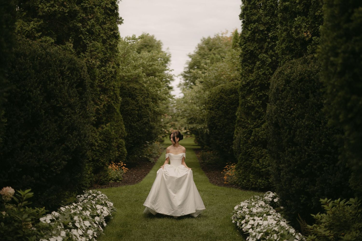 Bride in full ball gown walks through a symmetrical garden path flanked by tall hedges and white flowers.