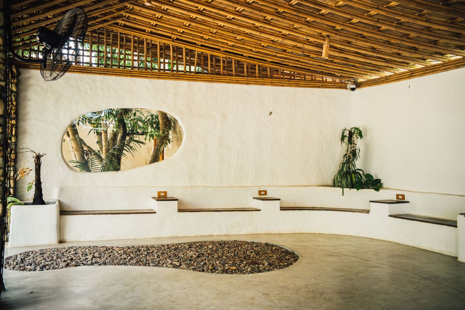 Serene open-air bamboo ceiling lounge with white curved walls, stone garden, tropical plants, and built-in white benches.