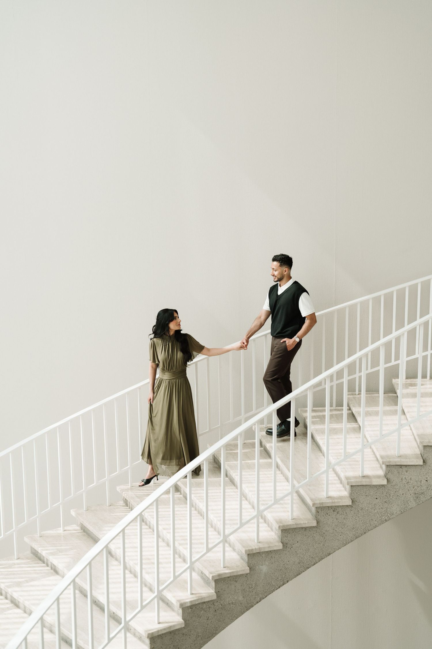 Couple holding hands on elegant white spiral staircase, woman in olive dress, man in dark vest, modern interior.