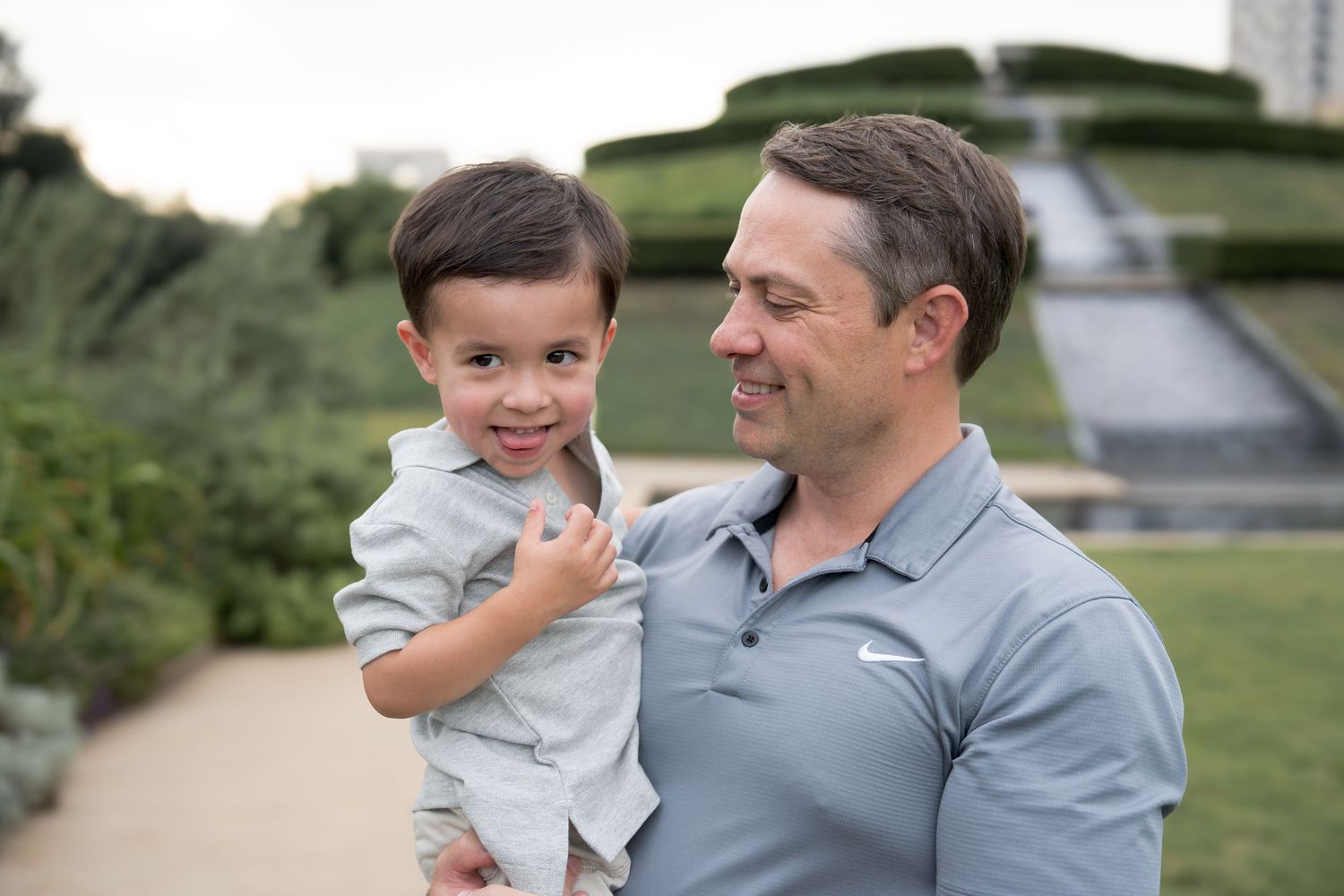 A smiling man in a gray Nike polo holds a young toddler boy outdoors near a fountain in a garden.
