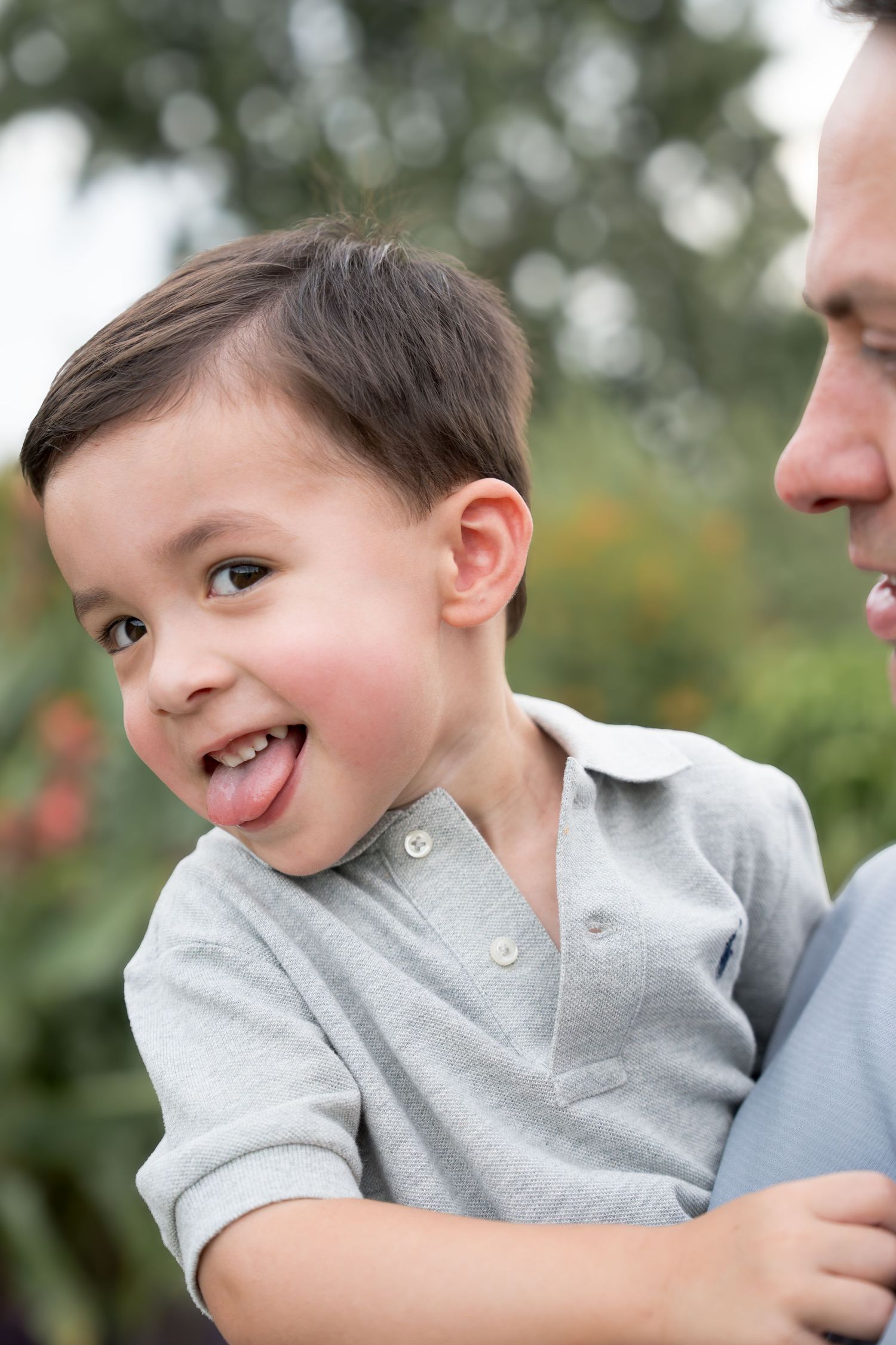 A playful toddler boy sticking his tongue out while being held by his father outdoors.