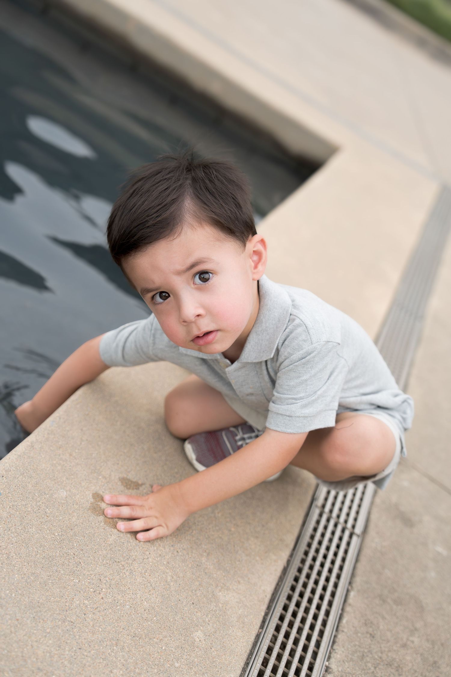 A young boy in a gray polo shirt crouching by the edge of a swimming pool.