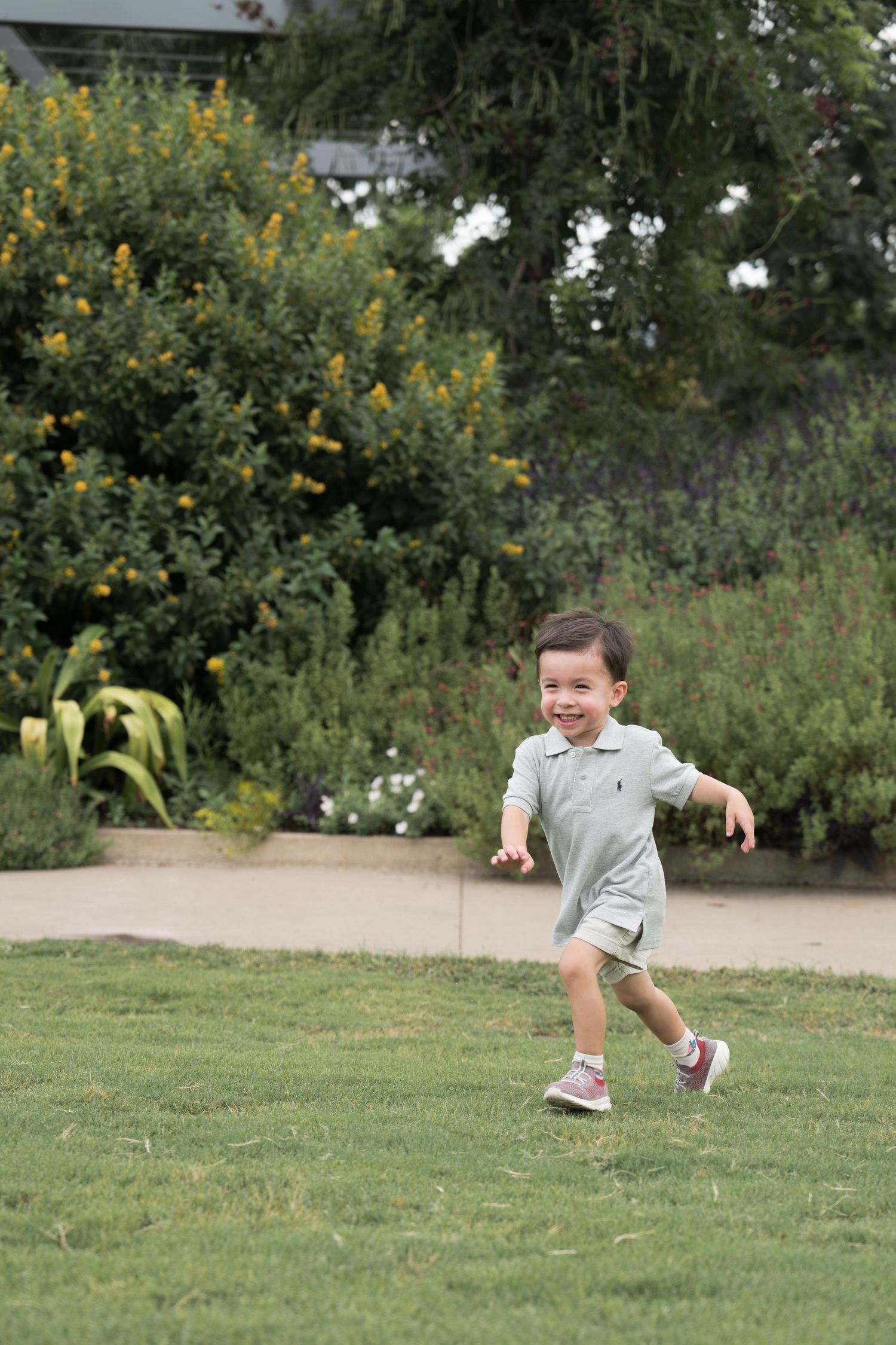 A happy toddler boy in a gray romper runs across green grass in a park with colorful flowers behind him.