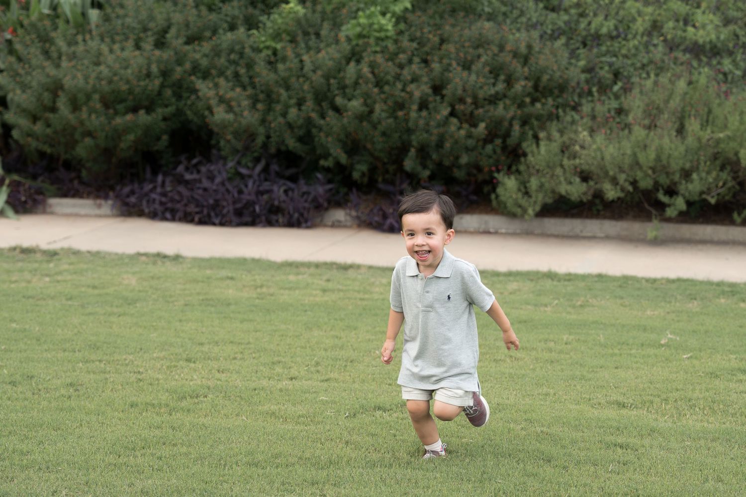 Happy toddler boy running on green grass lawn in a park with trees and shrubs in the background.