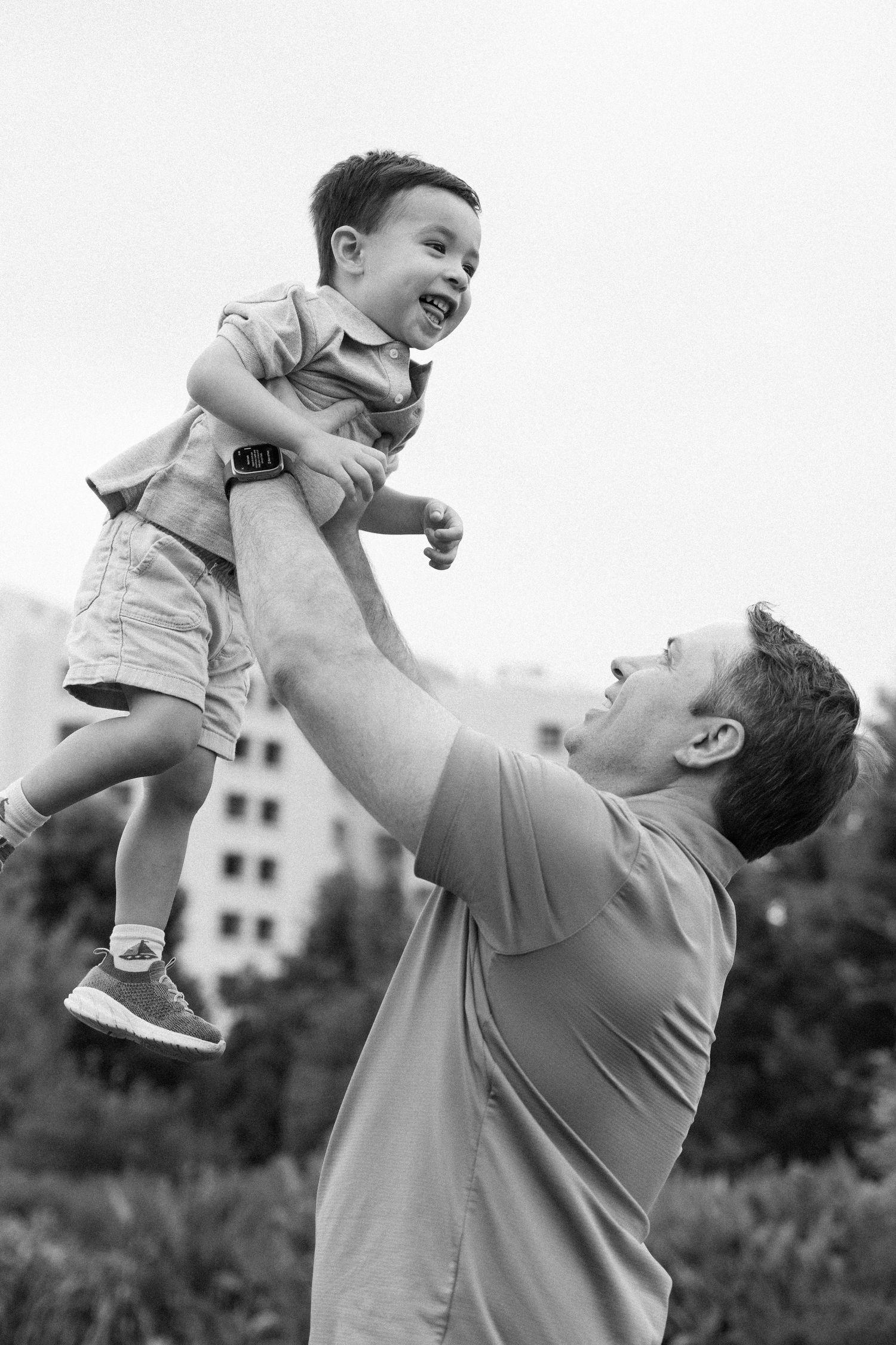 Black and white photo of a joyful father lifting his laughing toddler son high in the air outdoors.