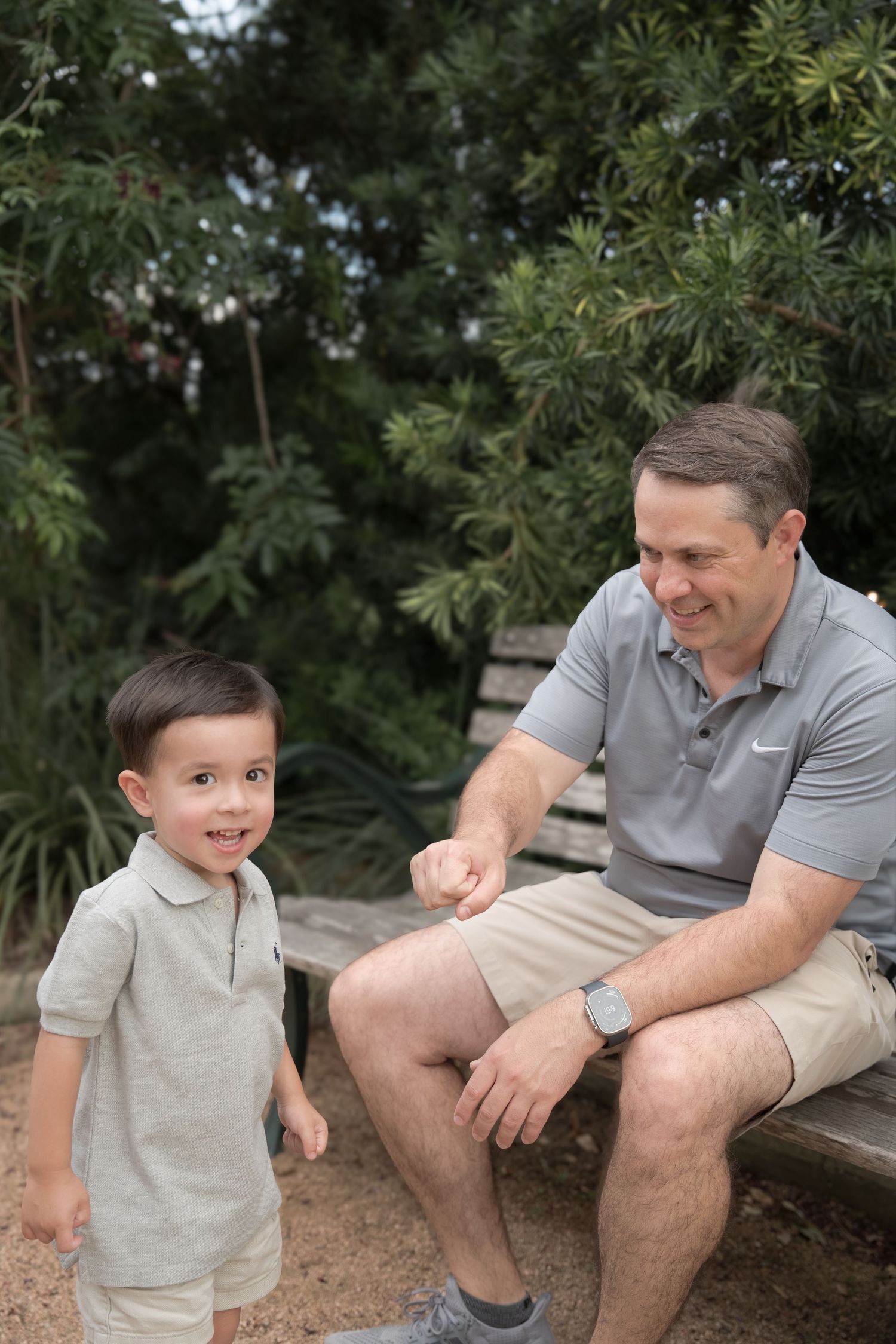 A smiling man in a gray Nike polo sits on a bench interacting with a happy young boy outdoors.