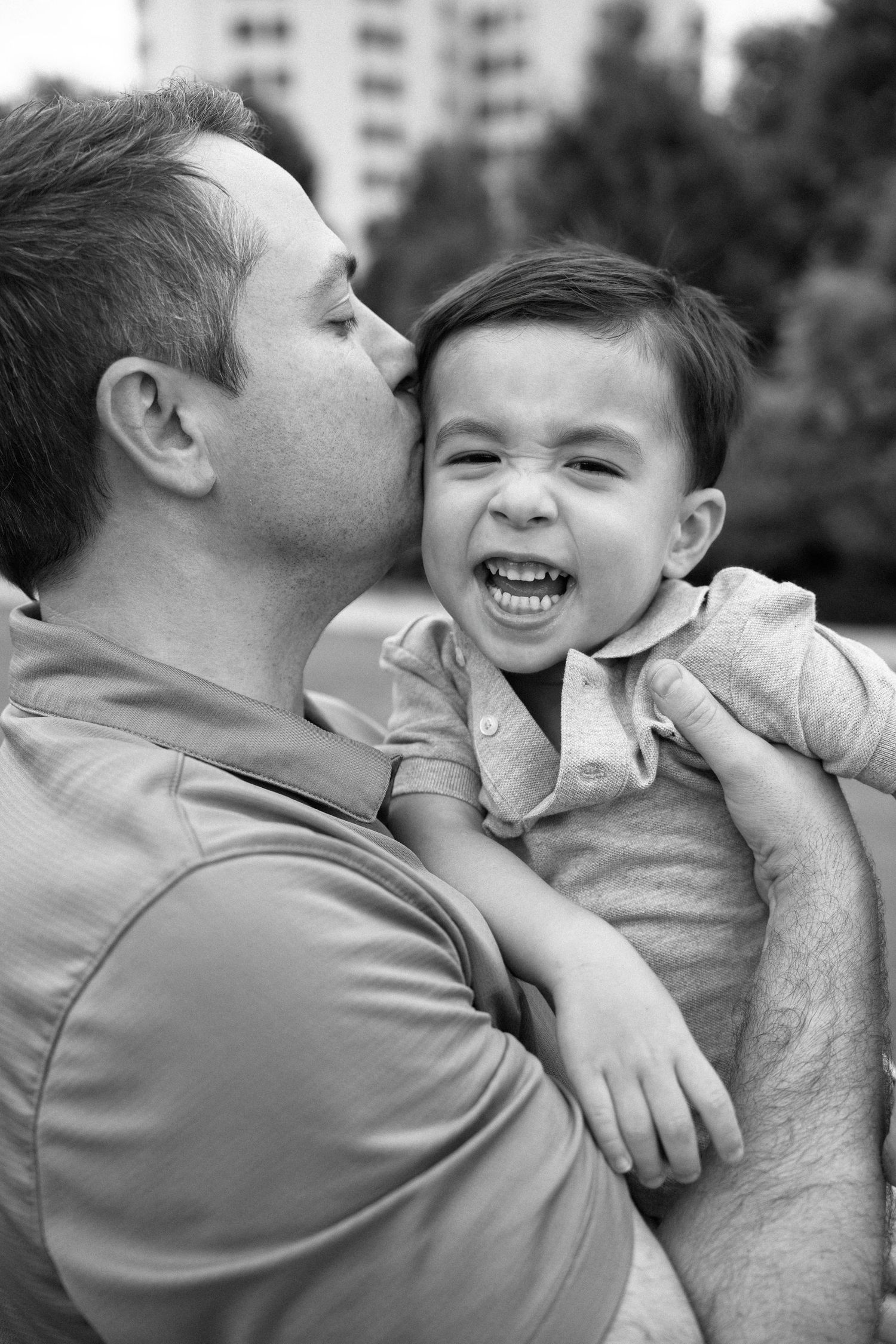 Black and white photo of a father kissing his laughing toddler son outdoors near trees and buildings.