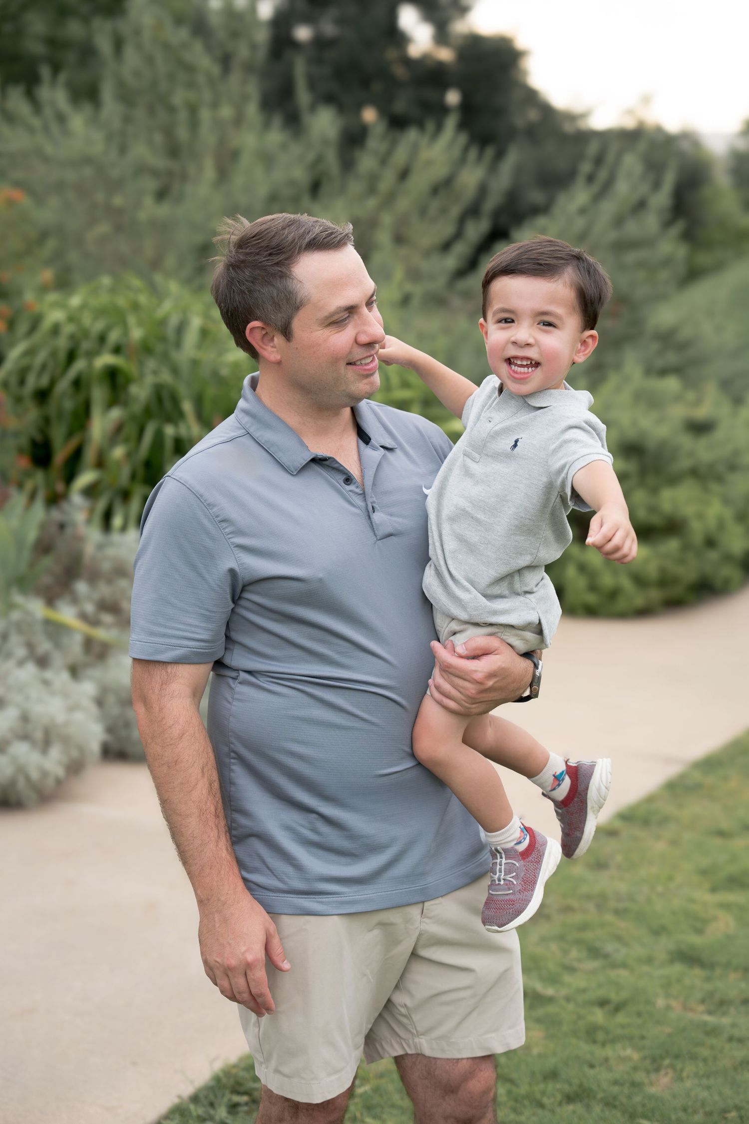 A smiling father holds his happy toddler son outdoors in a lush garden setting, both wearing matching gray polo shirts.