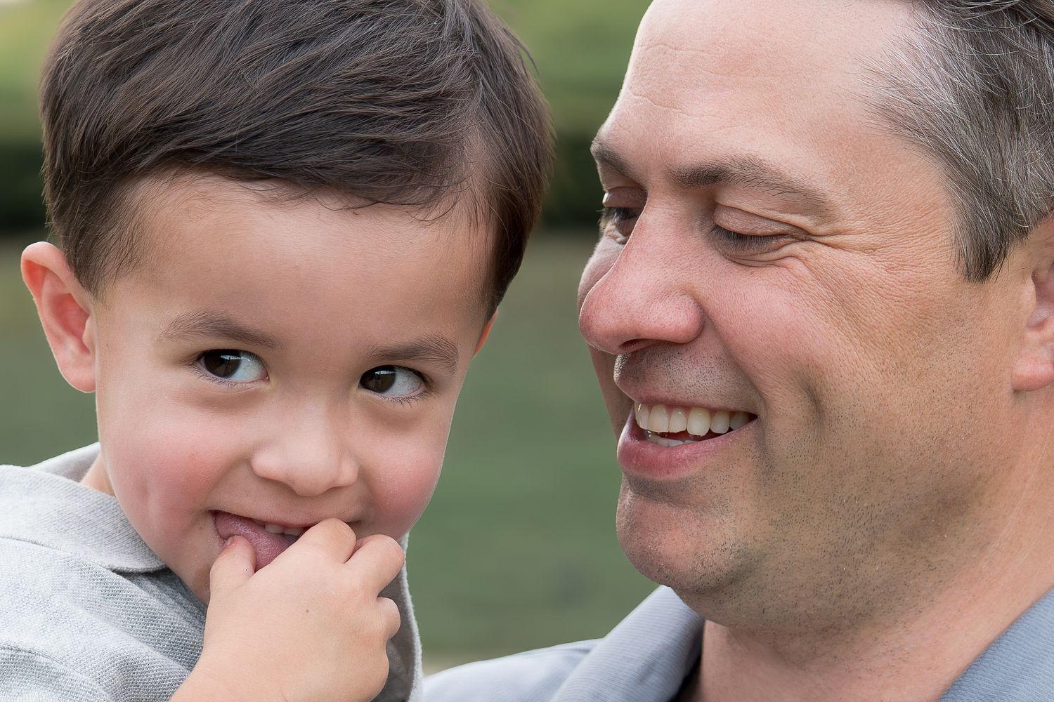 A smiling father holds his young son close, sharing a joyful moment outdoors in a park setting.