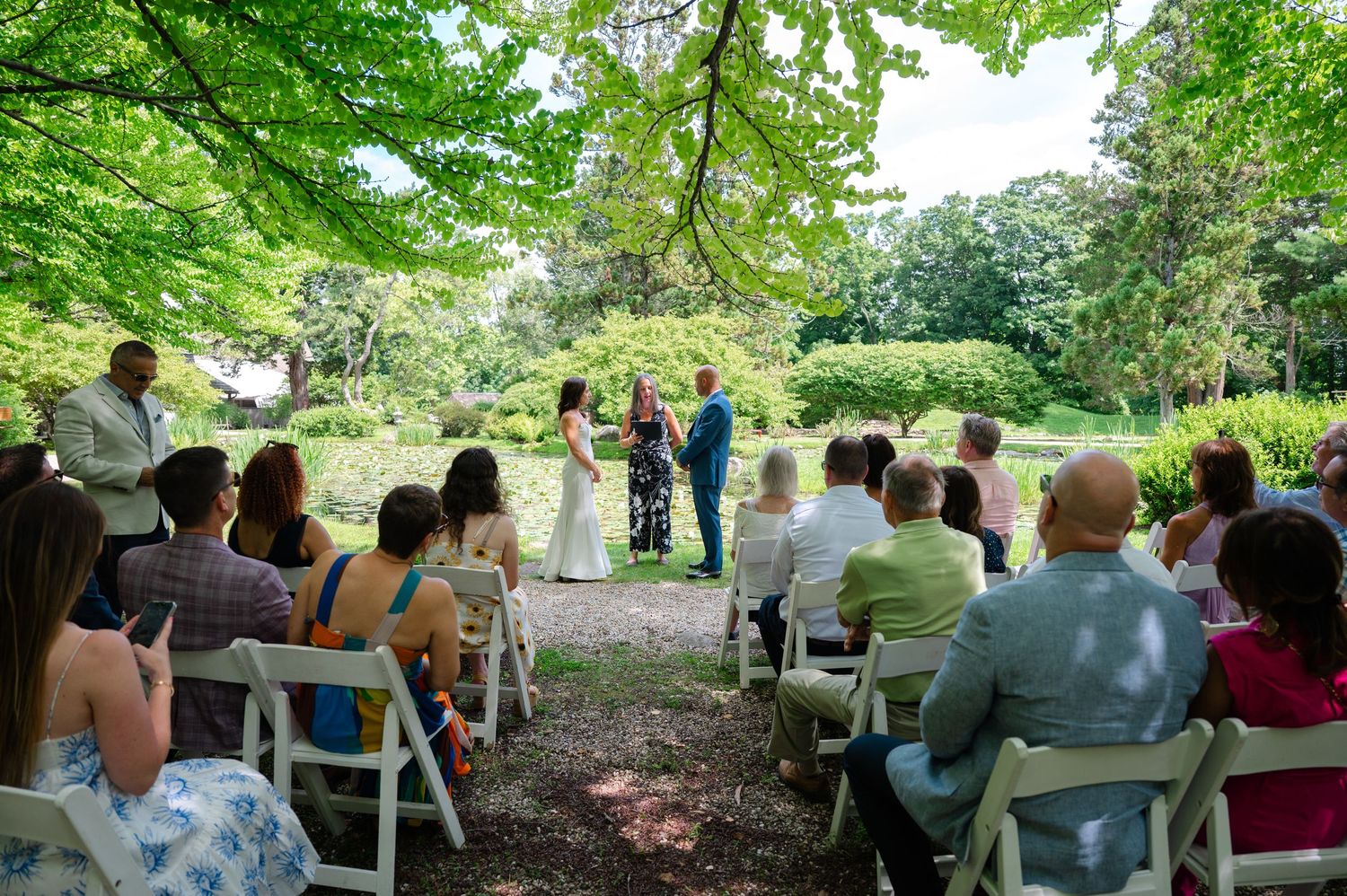 Outdoor wedding ceremony in Japanese Stroll Garden with guests seated on white chairs watching couple exchange vows in North Salem, NY.