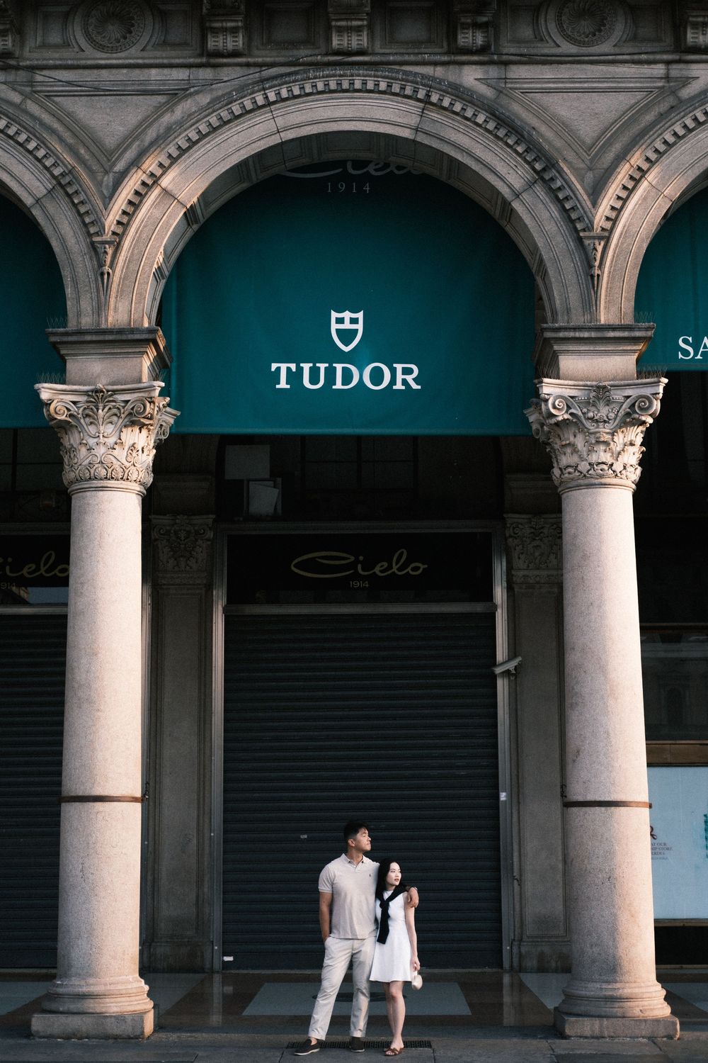 Couple standing under stone arches in front of a Tudor watch store with teal awning in an elegant European arcade.
