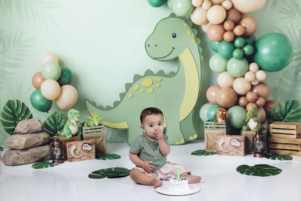 Baby boy sits before a cake at a dinosaur-themed first birthday party with green balloons and tropical decor.